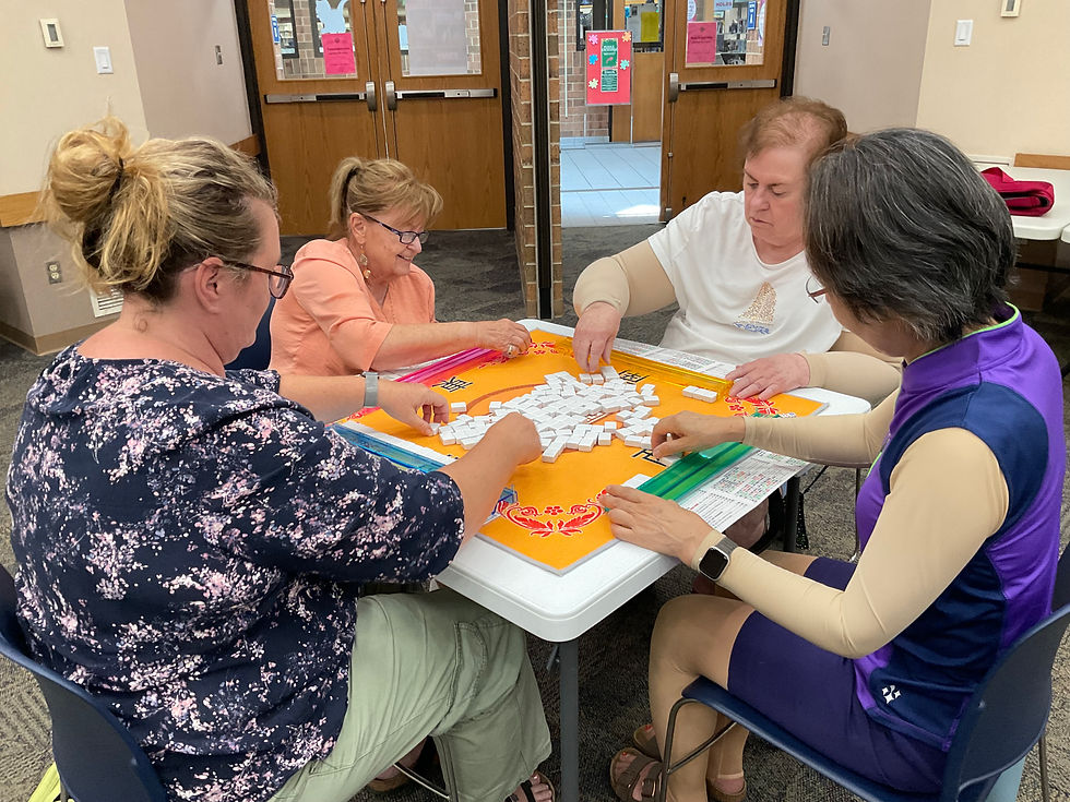 Four people sit at a square table with a mahjong board arranging face down white tiles