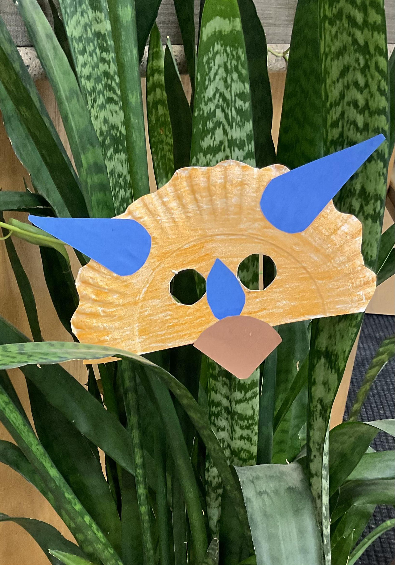 A triceratops mask made of half a paper plate with eye holes cut in and paper horns, nose, and beak added
