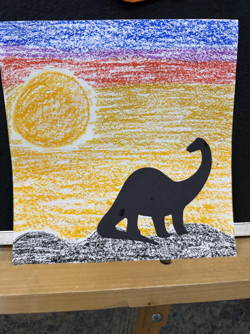 The finished activity shows a sunset drawn on a sheet of paper, with the paper dinosaur cut out pasted in the foreground