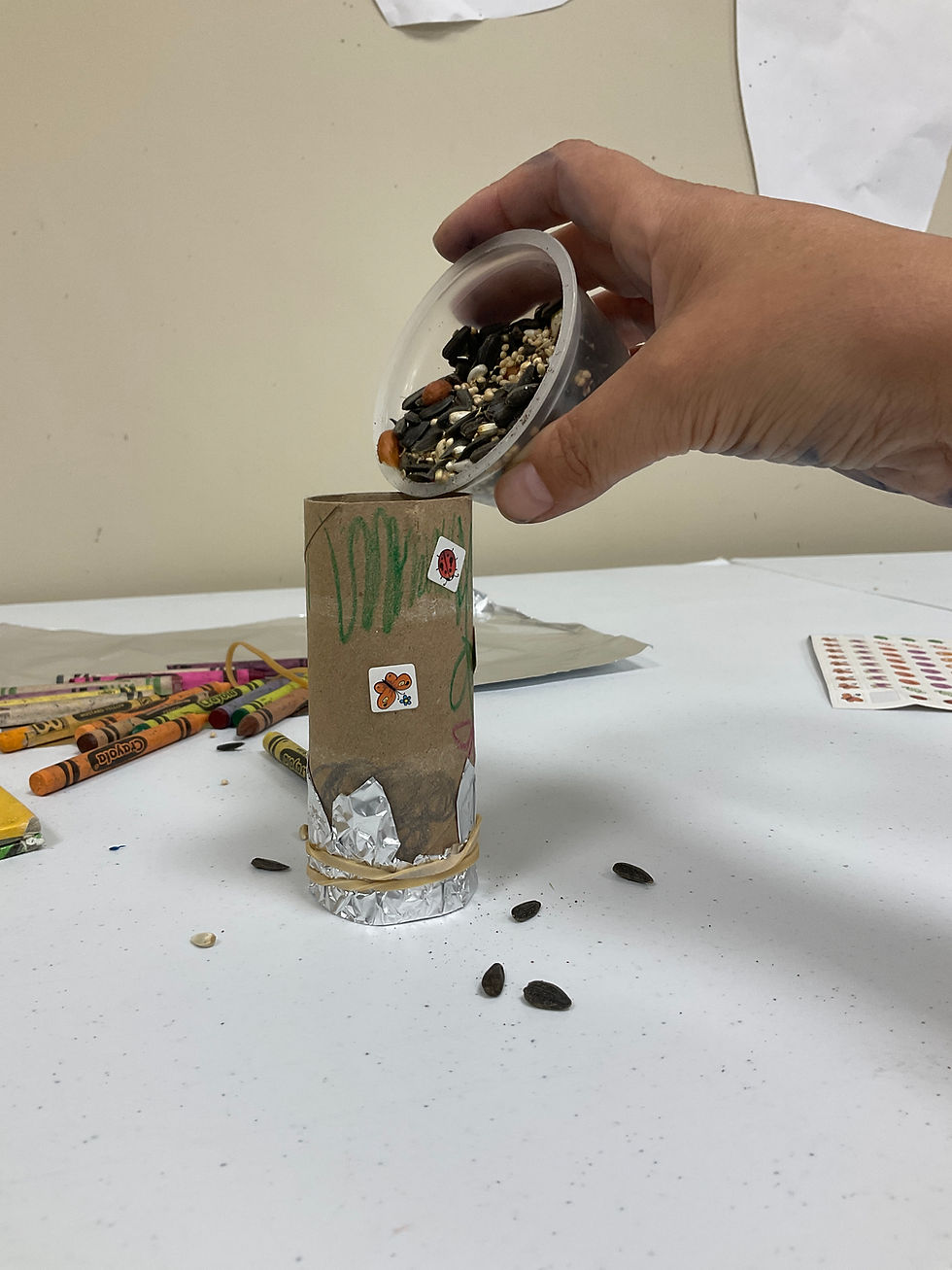 A hand pours seeds into a cardboard tube with one end covered by a foil cap