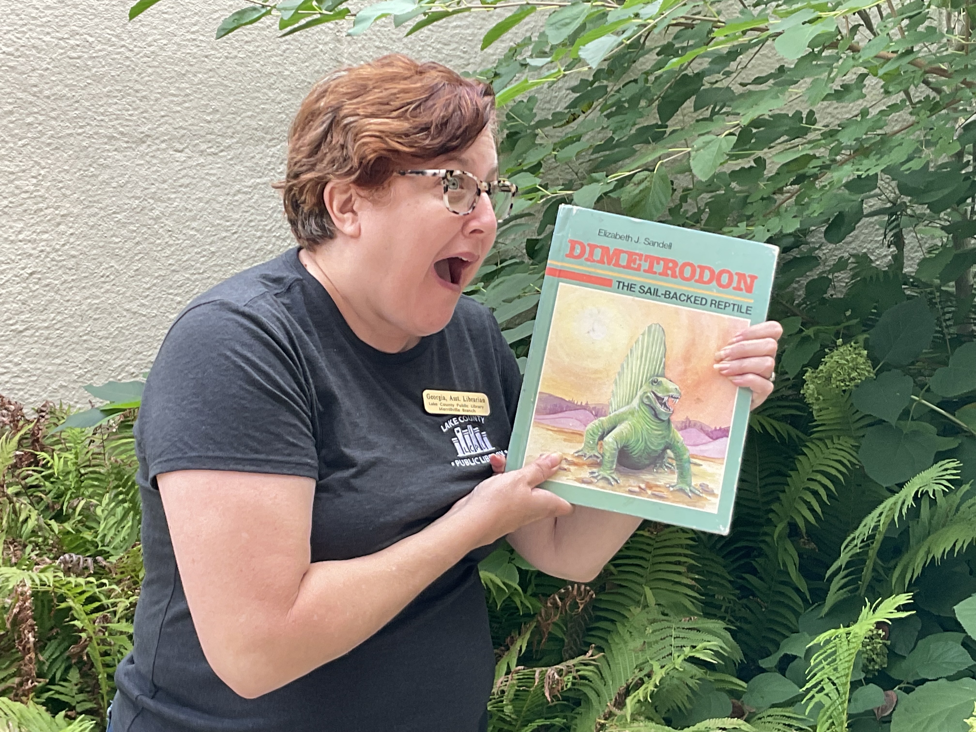 A library staff member holds up an old book about Dimetrodon and mimics the reptile's open mouthed grinning expression