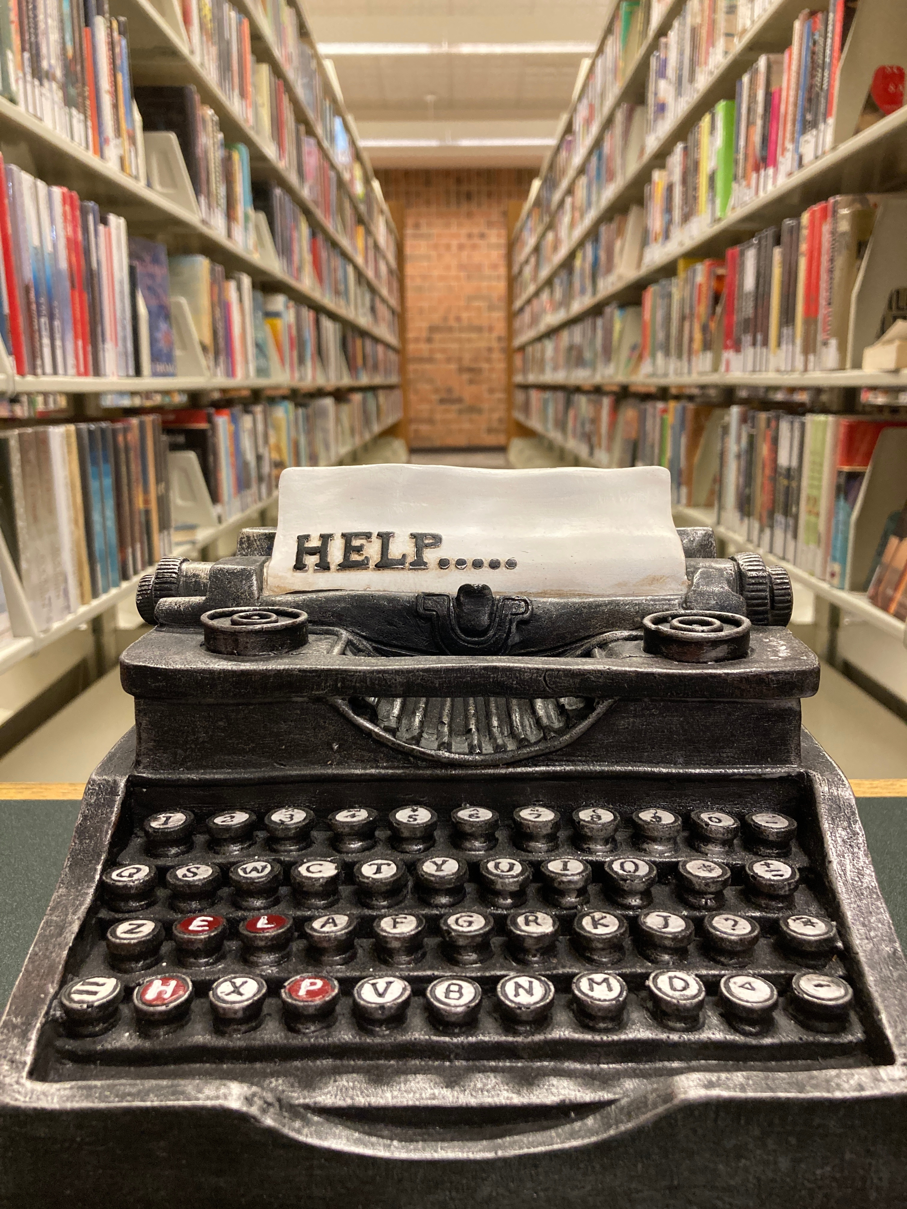 A small typewriter paperweight. The paper in it says Help. Stacks of books stretch out behind it