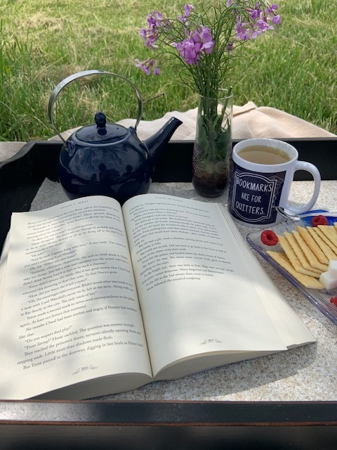 An open book sitting on a tray in the grass with a vase of purple flowers, a teapot, a cracker and cheese plate, and a mug that says bookmarks are for quitters