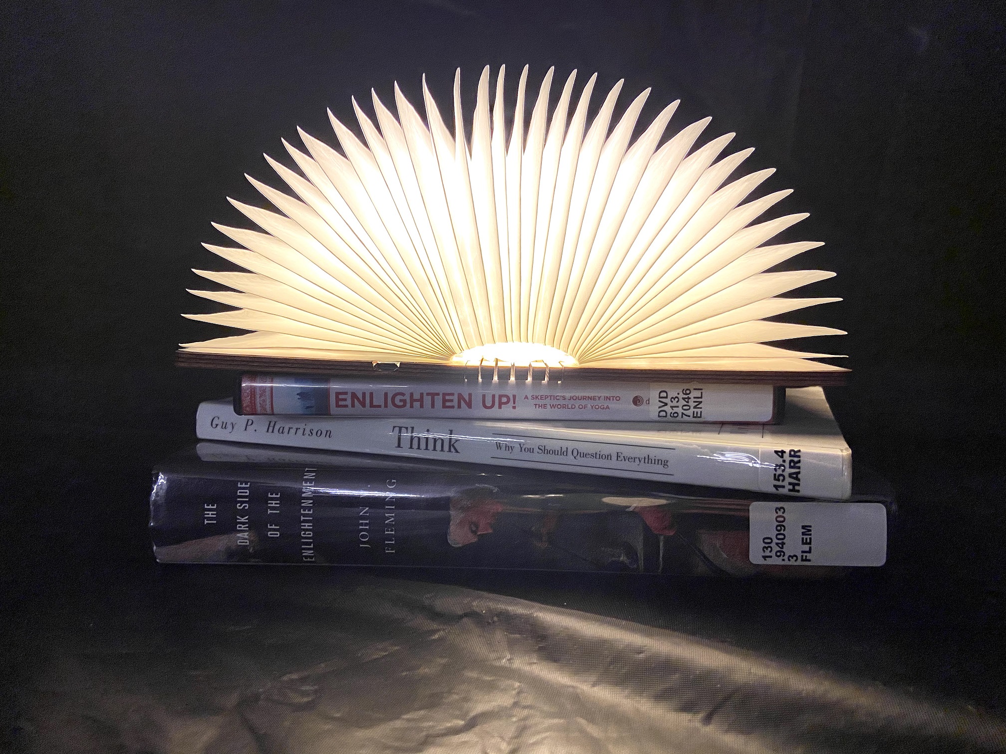 A lamp in the shape of an open book, its pages glowing. It sits on top of a DVD called Enlighten Up, a book called Think by Guy P Harrison, and a book called The Dark Side of the Enlightenment by John Fleming