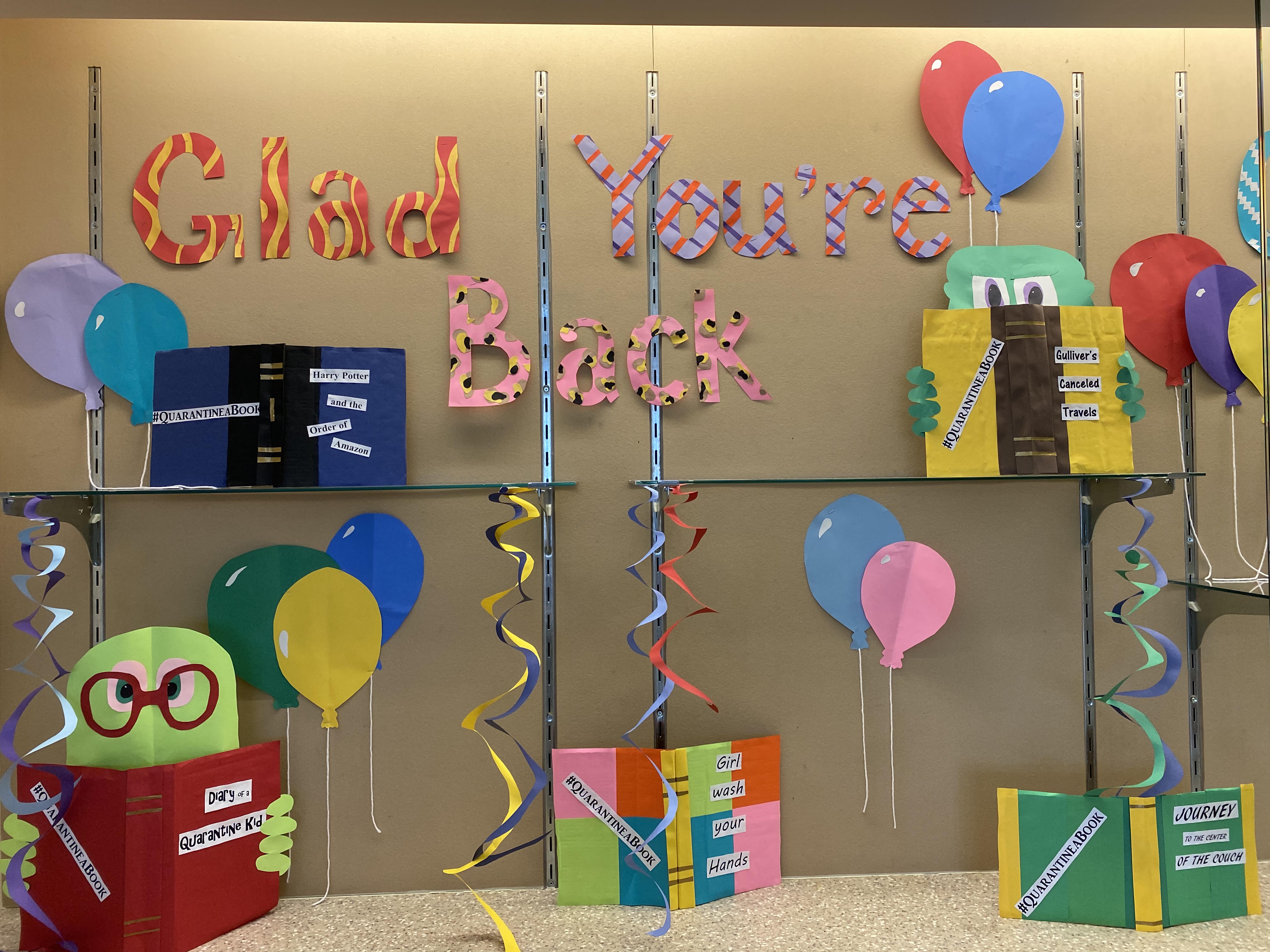 Display case filled with paper cut out decorations: Balloons, letters spelling Glad You're Back, bookworms, and books