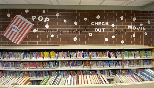 The brick wall above a short bookshelf is decorated with a paper cut out of a large popcorn bag and scattered popcorn pieces. Letters spell out Pop into the library and check out a good movie