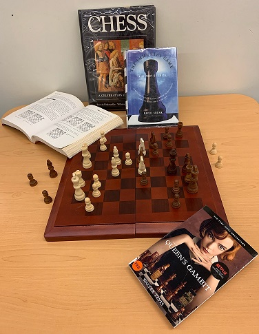 A chess board mid-game with chess instructional books stood up behind the board, one open on the corner. The novel The Queen's Gambit with the Netflix adaptation cover is in the foreground