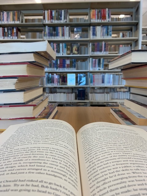 A book lying open on a table with two stacks of books in front of it, and beyond that library shelves filled with books