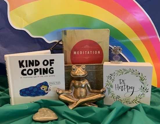 A bronze frog sitting cross legged under a rainbow with books on meditation, coping, and being happy